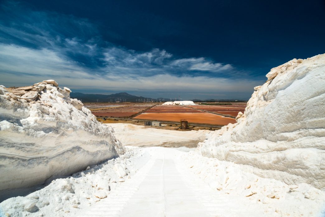 Veduta delle Saline Conti Vecchi, sale in primo piano, vasche, impianti, sullo sfondo pale eoliche e montagne, cielo azzurro