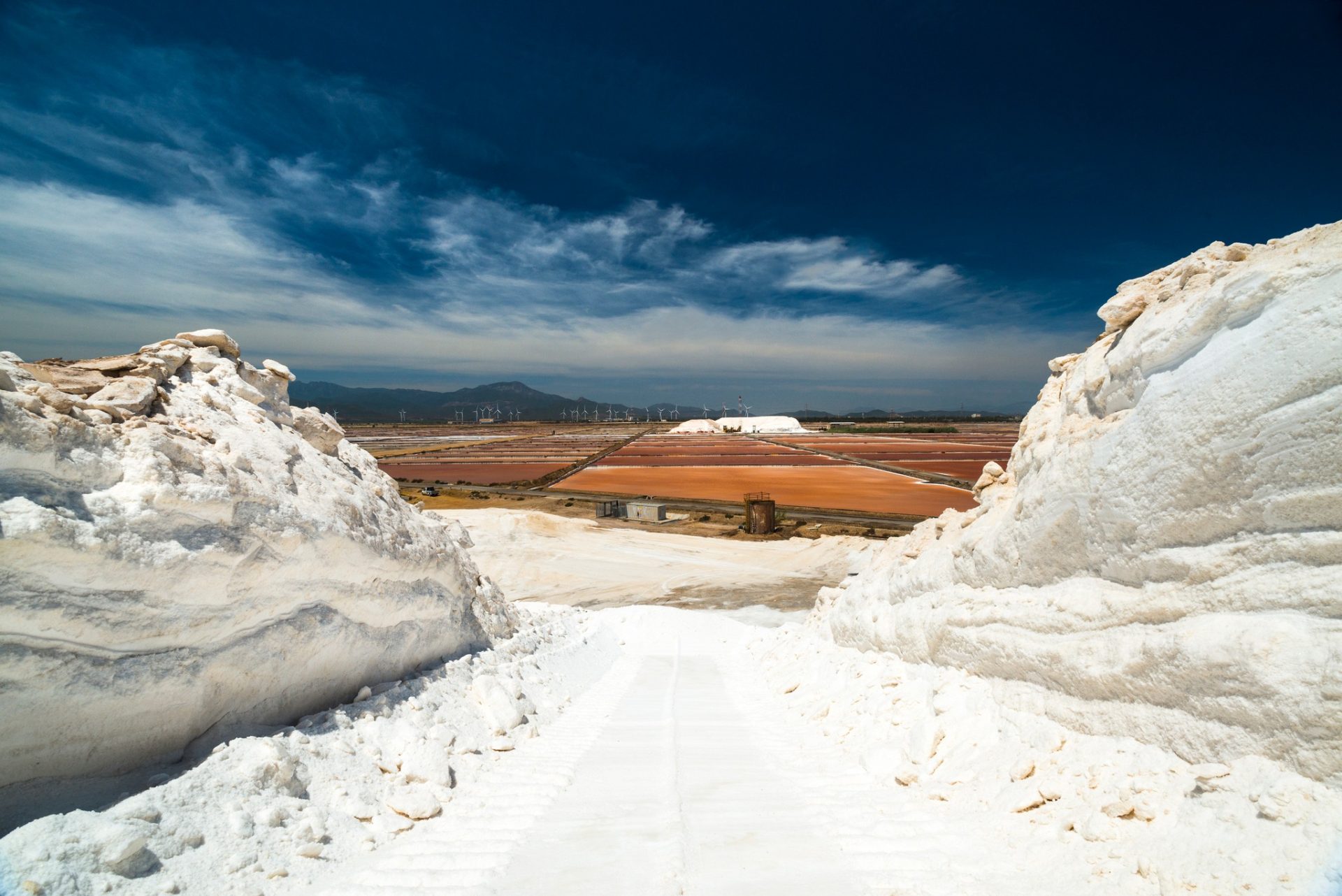 Veduta delle Saline Conti Vecchi, sale in primo piano, vasche, impianti, sullo sfondo pale eoliche e montagne, cielo azzurro