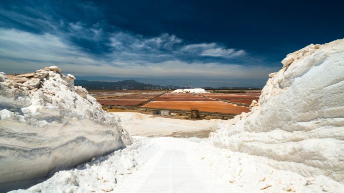 Veduta delle Saline Conti Vecchi, sale in primo piano, vasche, impianti, sullo sfondo pale eoliche e montagne, cielo azzurro